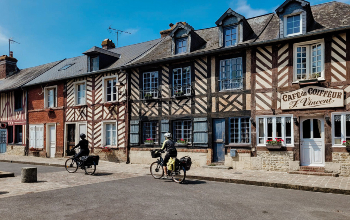 Scène paisible à Beuvron-en-Auge avec des cyclistes passant devant des maisons à colombages traditionnelles et un ancien café.