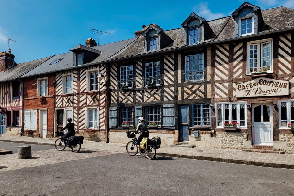 Scène paisible à Beuvron-en-Auge avec des cyclistes passant devant des maisons à colombages traditionnelles et un ancien café.